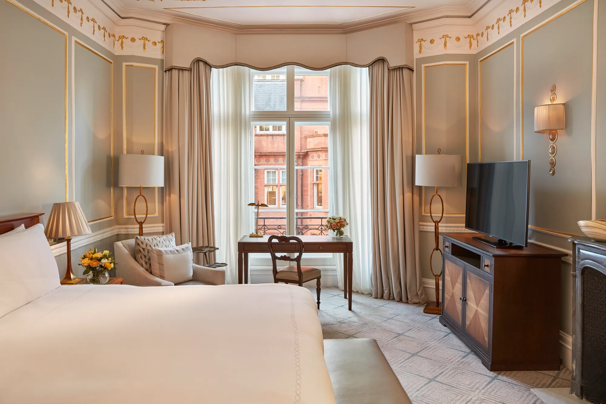 Elegant bedroom with a large bed, writing desk by a tall window, and classic décor featuring pale blue walls with gold trim and traditional furnishings.