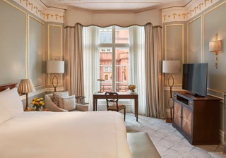 Elegant bedroom with a large bed, writing desk by a tall window, and classic décor featuring pale blue walls with gold trim and traditional furnishings.