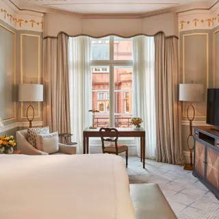 Elegant bedroom with a large bed, writing desk by a tall window, and classic décor featuring pale blue walls with gold trim and traditional furnishings.