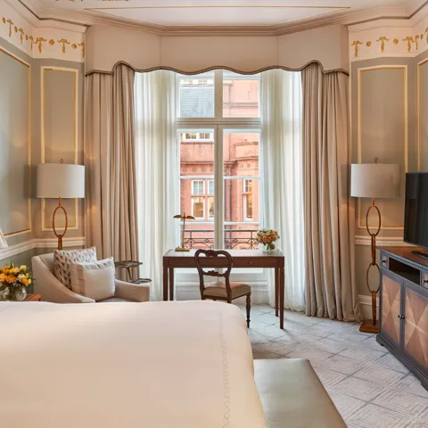 Elegant bedroom with a large bed, writing desk by a tall window, and classic décor featuring pale blue walls with gold trim and traditional furnishings.