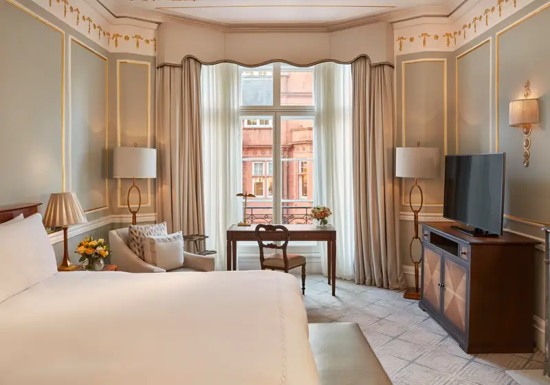 Elegant bedroom with a large bed, writing desk by a tall window, and classic décor featuring pale blue walls with gold trim and traditional furnishings.