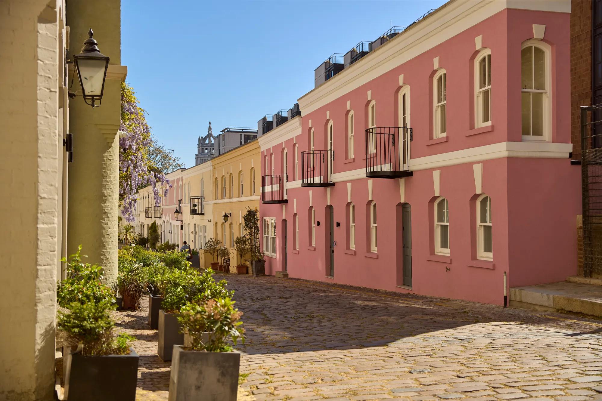 Pastel pink and cream townhouses lining a cobbled mews street, with potted plants and balconies under clear blue skies.