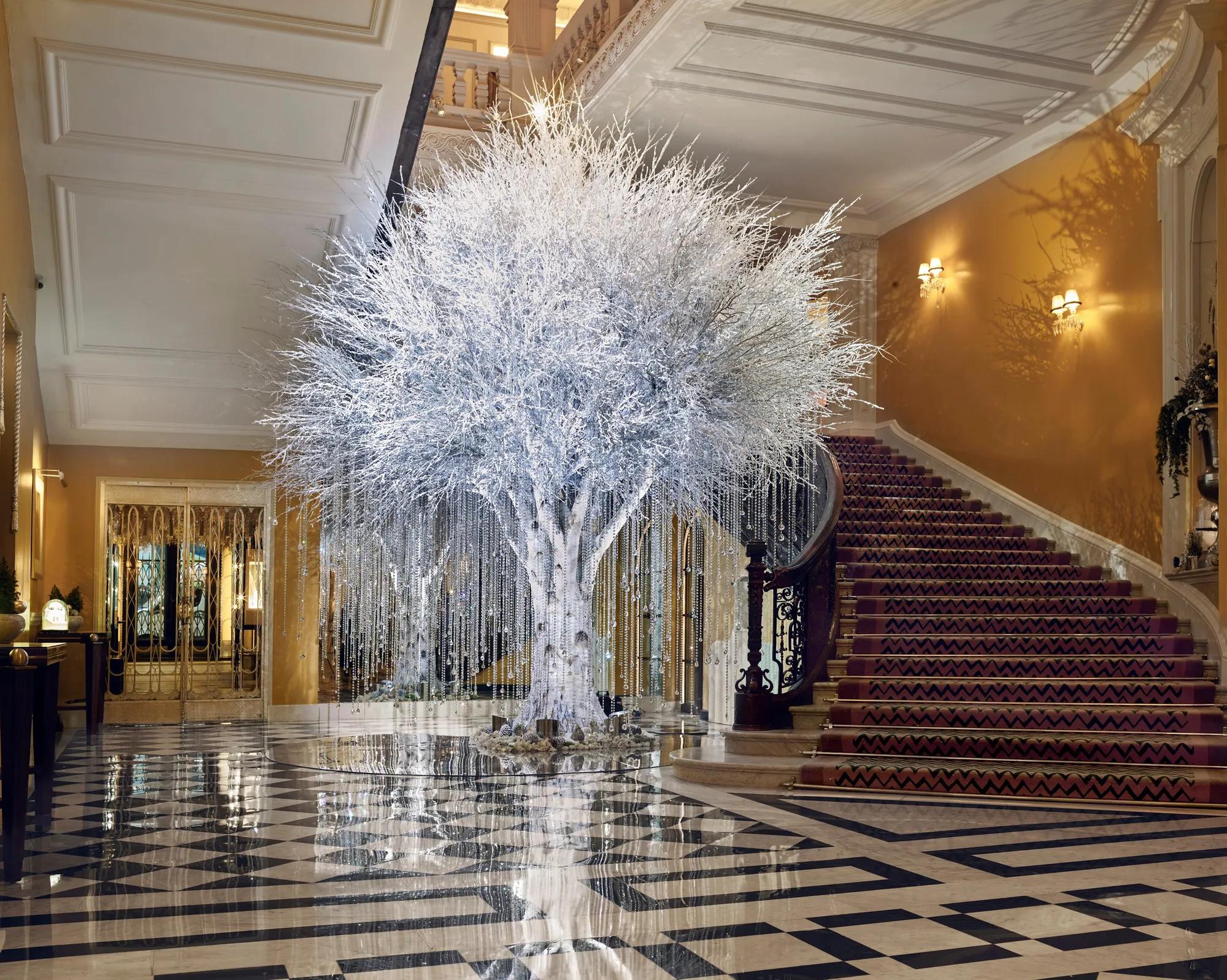Grand hotel lobby with sweeping staircase and checkered floor, featuring a white sculptural tree with hanging crystal strands reflecting light across the space.