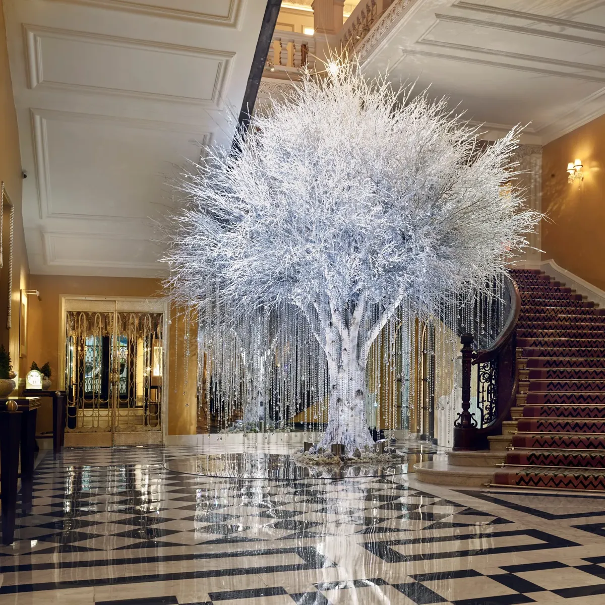 Grand hotel lobby with sweeping staircase and checkered floor, featuring a white sculptural tree with hanging crystal strands reflecting light across the space.