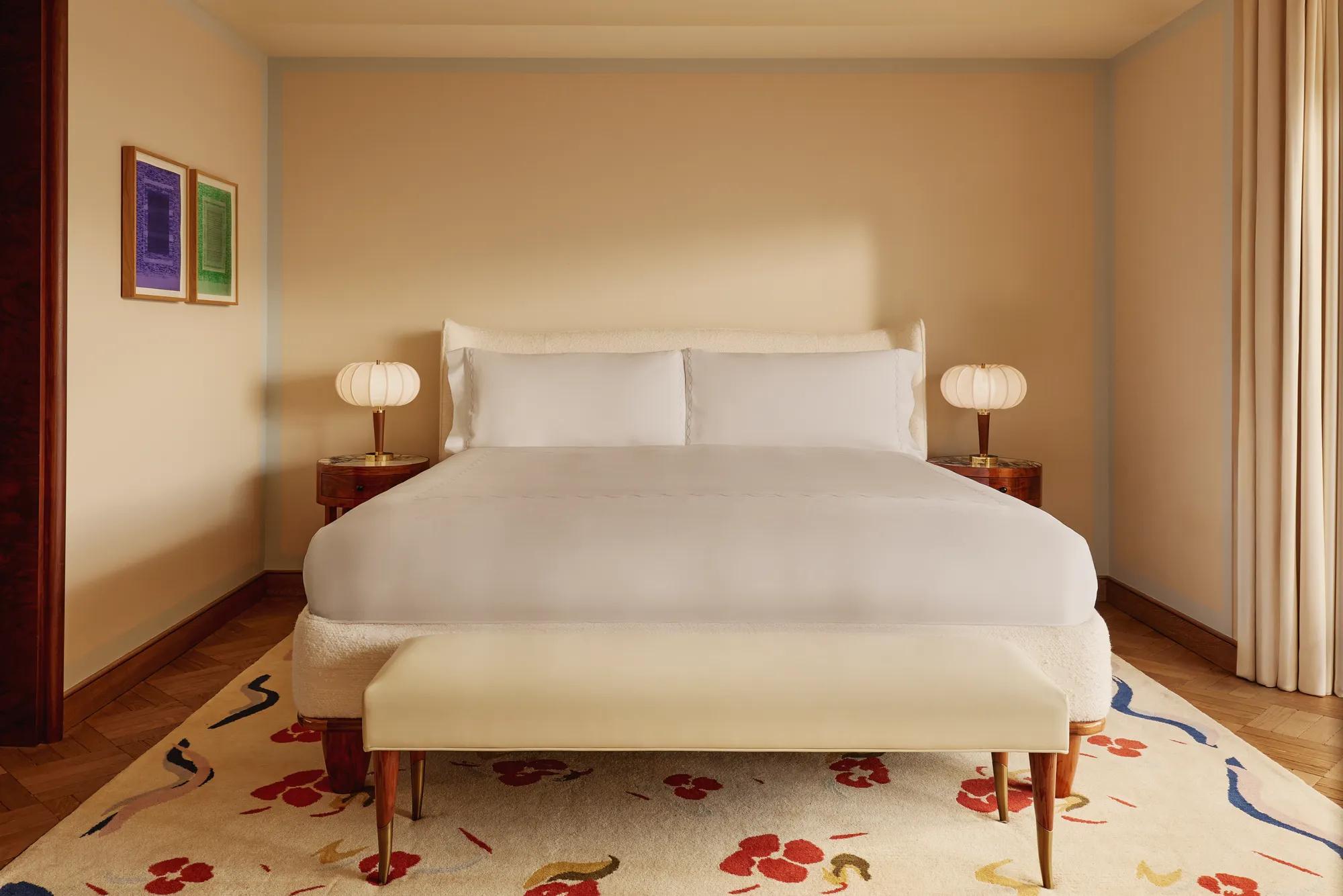 Minimalist bedroom with a king-size bed dressed in white linens, rounded bedside tables with sculptural lamps, a cream bench, and a patterned rug with red and blue floral accents.