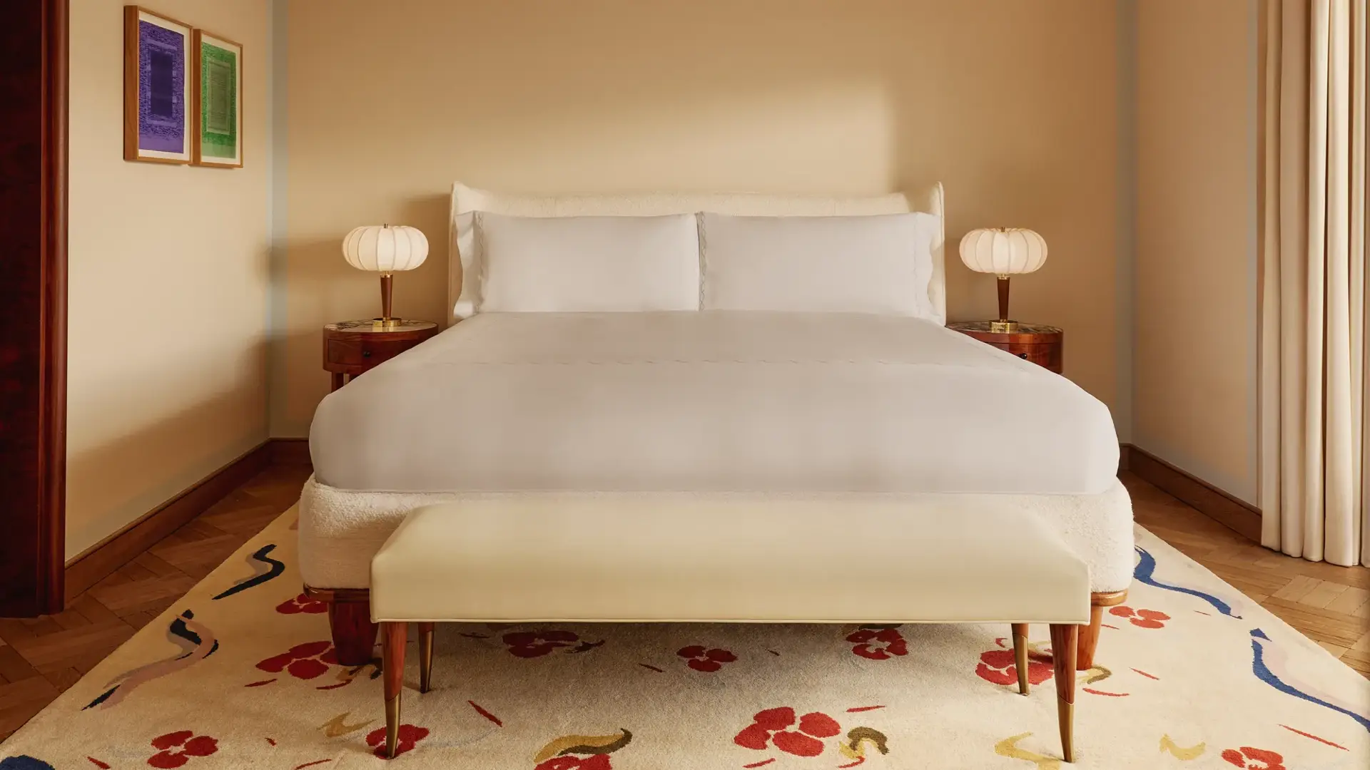 Minimalist bedroom with a king-size bed dressed in white linens, rounded bedside tables with sculptural lamps, a cream bench, and a patterned rug with red and blue floral accents.