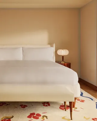 Minimalist bedroom with a king-size bed dressed in white linens, rounded bedside tables with sculptural lamps, a cream bench, and a patterned rug with red and blue floral accents.