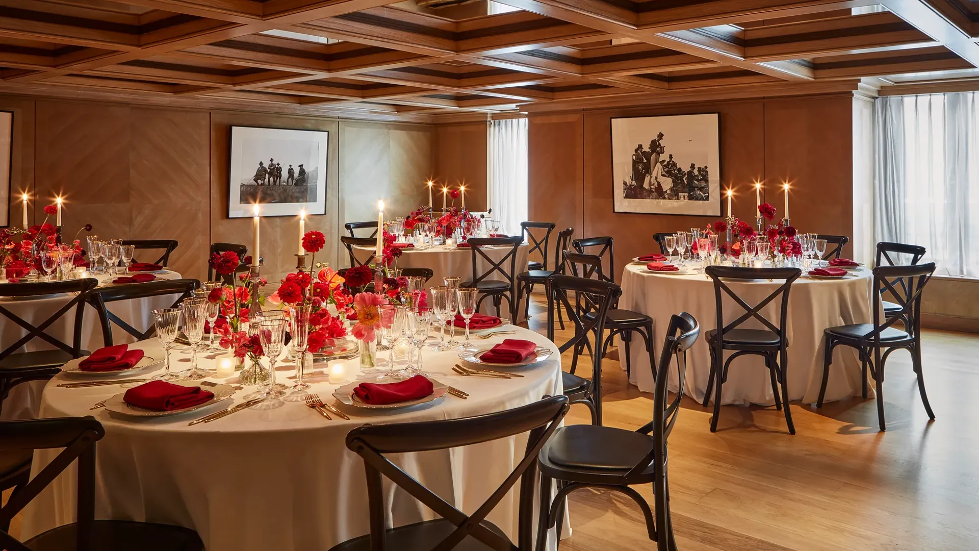 Elegant private dining room with round tables set for an event, decorated with red flowers, candles, and neatly folded red napkins on white tablecloths. The wood-panelled walls and coffered ceiling create a warm, intimate atmosphere, enhanced by natural light through sheer curtains.