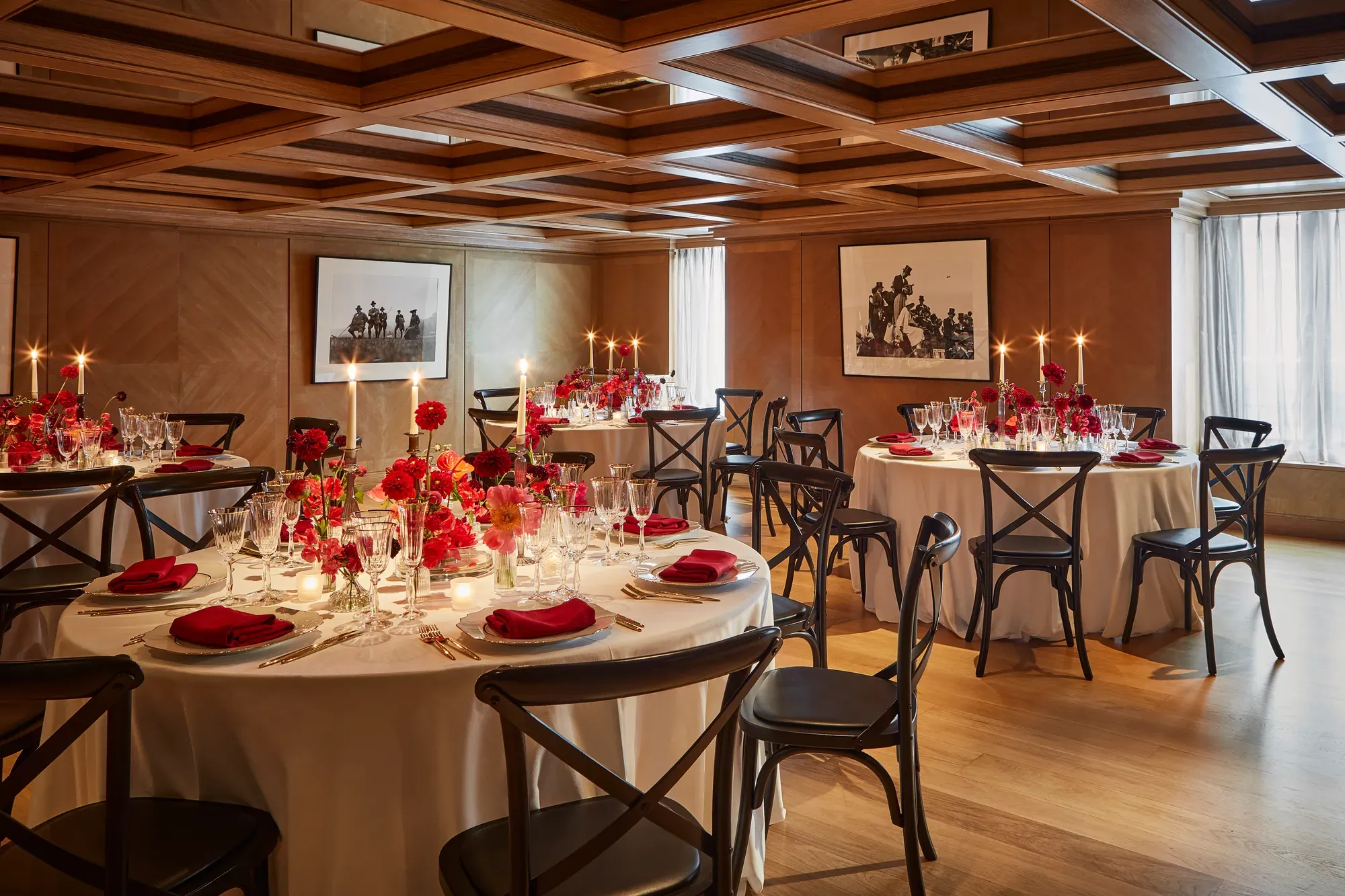 Elegant private dining room with round tables set for an event, decorated with red flowers, candles, and neatly folded red napkins on white tablecloths. The wood-panelled walls and coffered ceiling create a warm, intimate atmosphere, enhanced by natural light through sheer curtains.