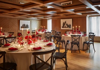 Elegant private dining room with round tables set for an event, decorated with red flowers, candles, and neatly folded red napkins on white tablecloths. The wood-panelled walls and coffered ceiling create a warm, intimate atmosphere, enhanced by natural light through sheer curtains.
