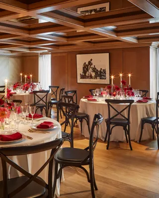 Elegant private dining room with round tables set for an event, decorated with red flowers, candles, and neatly folded red napkins on white tablecloths. The wood-panelled walls and coffered ceiling create a warm, intimate atmosphere, enhanced by natural light through sheer curtains.
