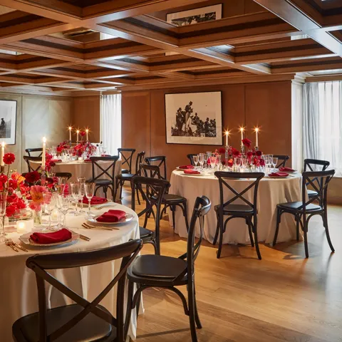 Elegant private dining room with round tables set for an event, decorated with red flowers, candles, and neatly folded red napkins on white tablecloths. The wood-panelled walls and coffered ceiling create a warm, intimate atmosphere, enhanced by natural light through sheer curtains.
