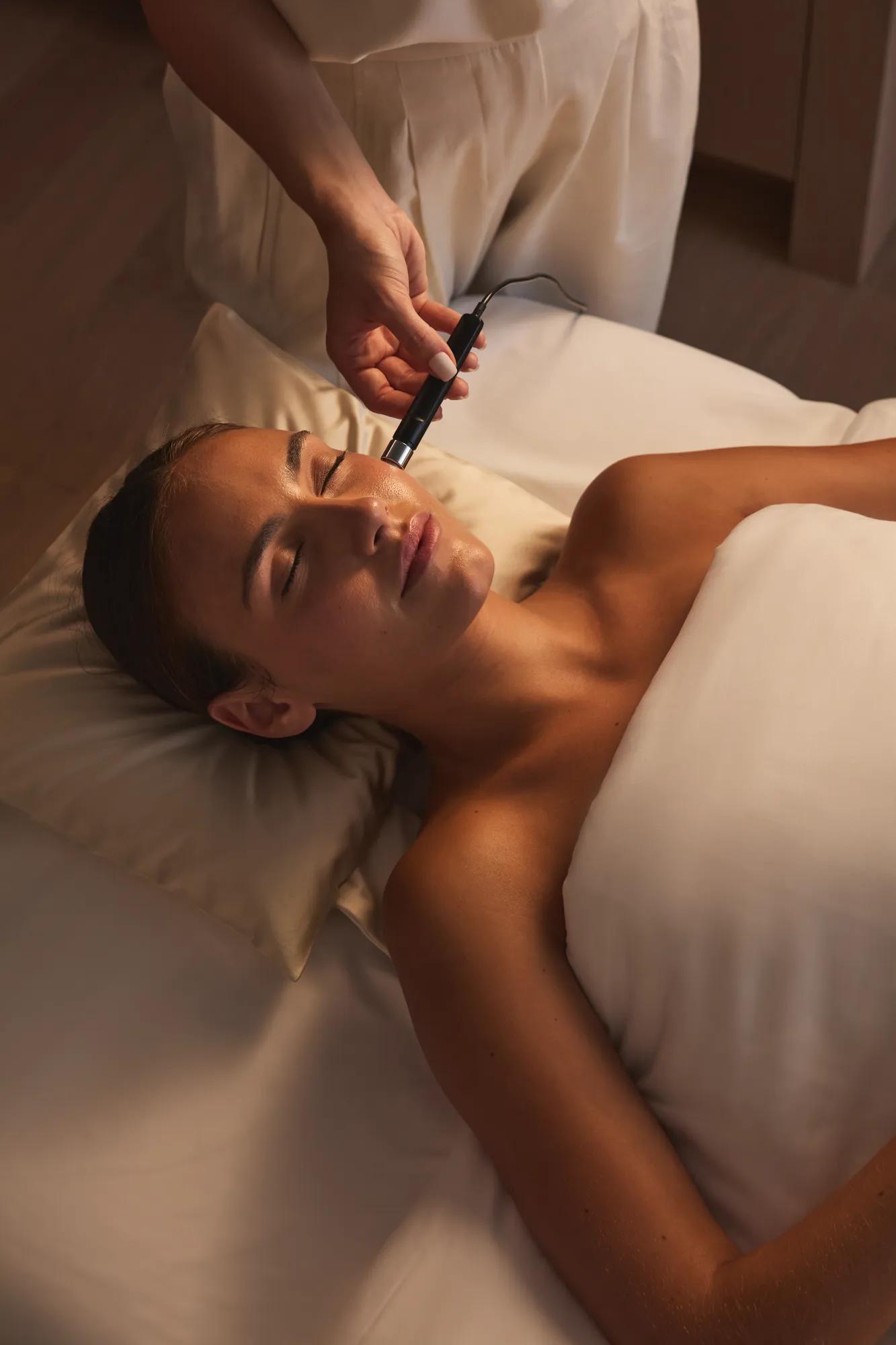 Spa therapist performing a gentle facial treatment using a handheld device in a calm, softly lit treatment room.