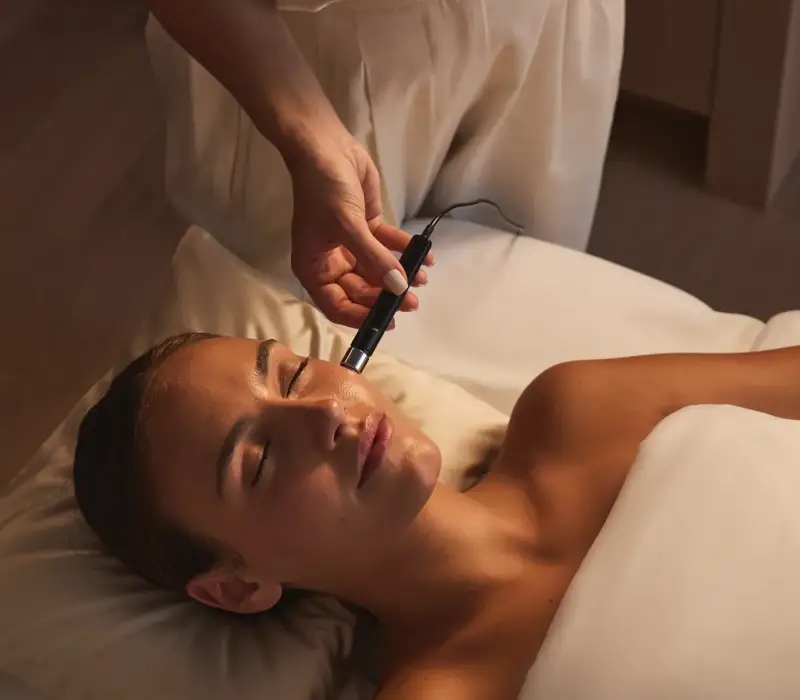 Spa therapist performing a gentle facial treatment using a handheld device in a calm, softly lit treatment room.