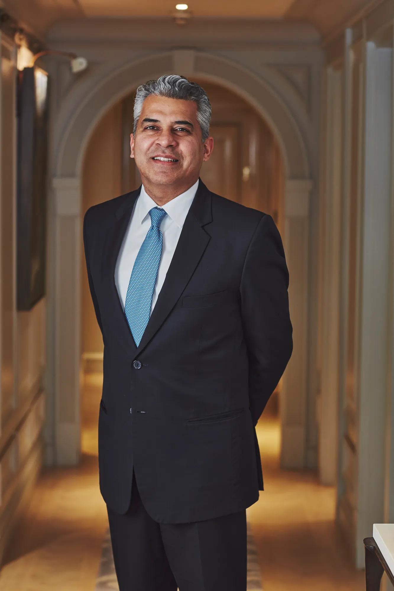 Portrait of a person in a dark suit and blue tie standing in an elegant hallway with arched architectural details.