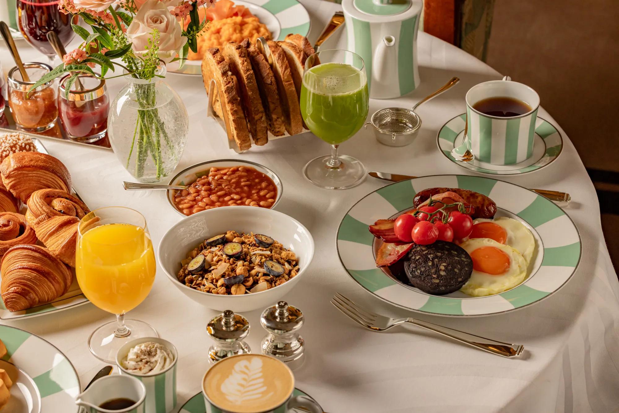 Breakfast table with cooked eggs, pastries, cereal, toast, fruit juices, and tea arranged on striped china and a white tablecloth