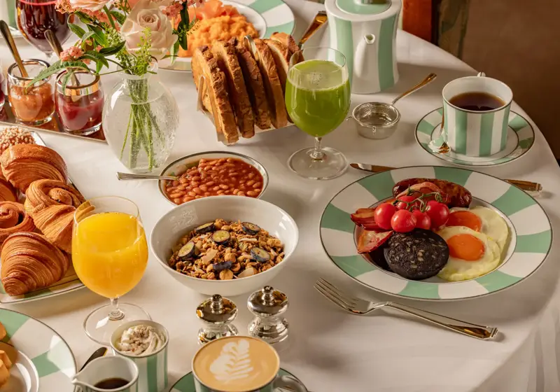 Breakfast table with cooked eggs, pastries, cereal, toast, fruit juices, and tea arranged on striped china and a white tablecloth