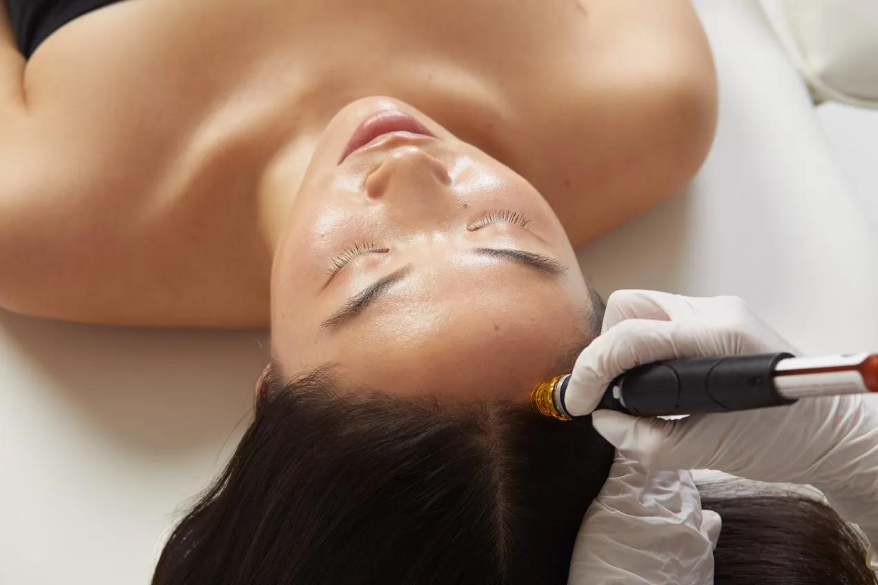 Woman receiving scalp treatment with handheld device, eyes closed, lying on treatment bed