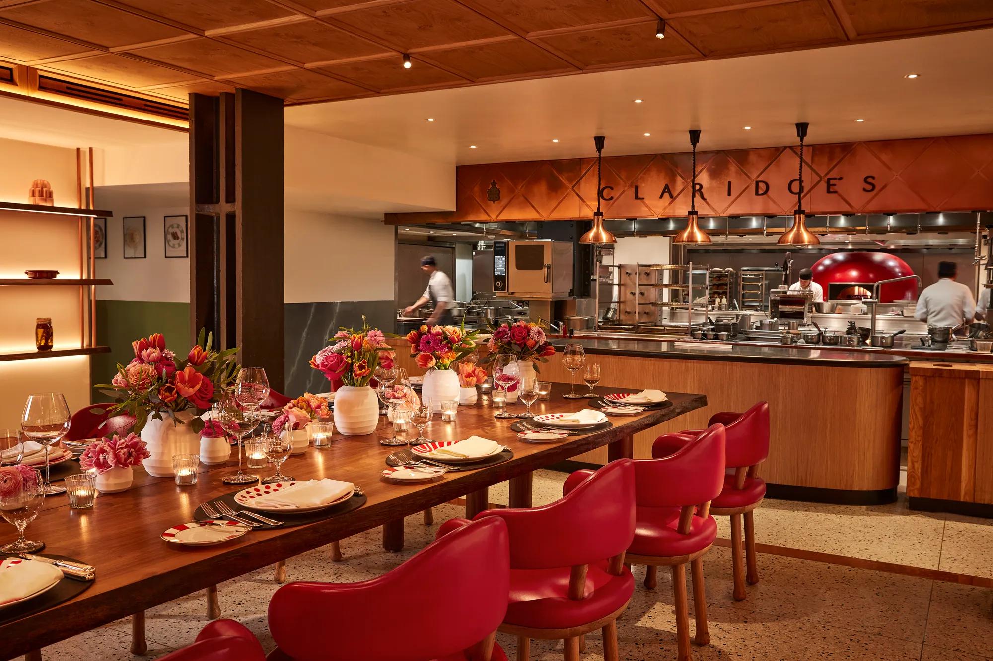 Warm, contemporary dining space with a long wooden table set with flowers, candles, and red chairs. The open kitchen in the background features copper accents, chefs at work, and “Claridge’s” displayed above the cooking area.