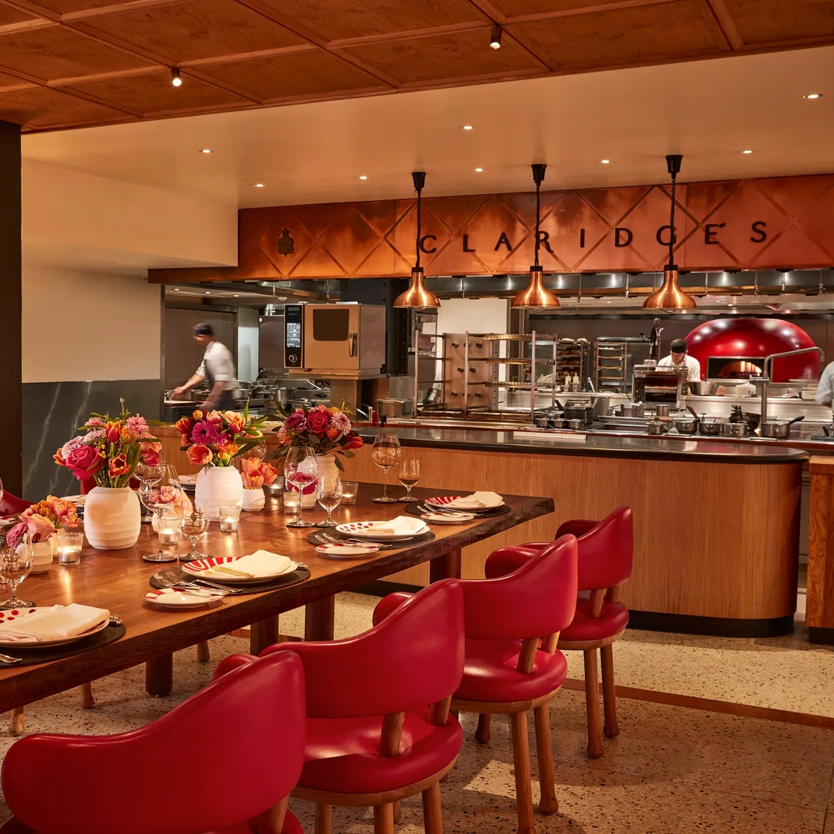 Warm, contemporary dining space with a long wooden table set with flowers, candles, and red chairs. The open kitchen in the background features copper accents, chefs at work, and “Claridge’s” displayed above the cooking area.