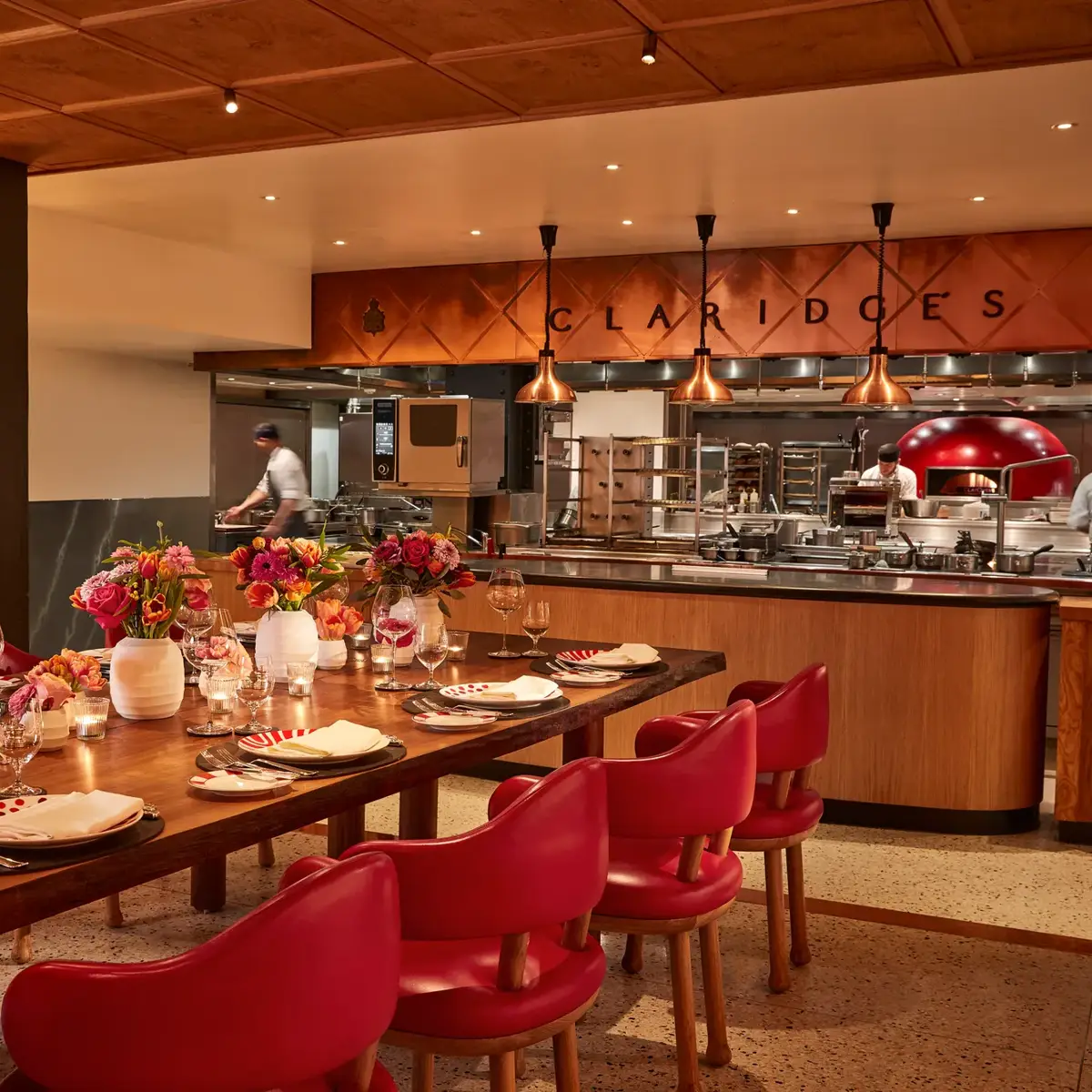 Warm, contemporary dining space with a long wooden table set with flowers, candles, and red chairs. The open kitchen in the background features copper accents, chefs at work, and “Claridge’s” displayed above the cooking area.