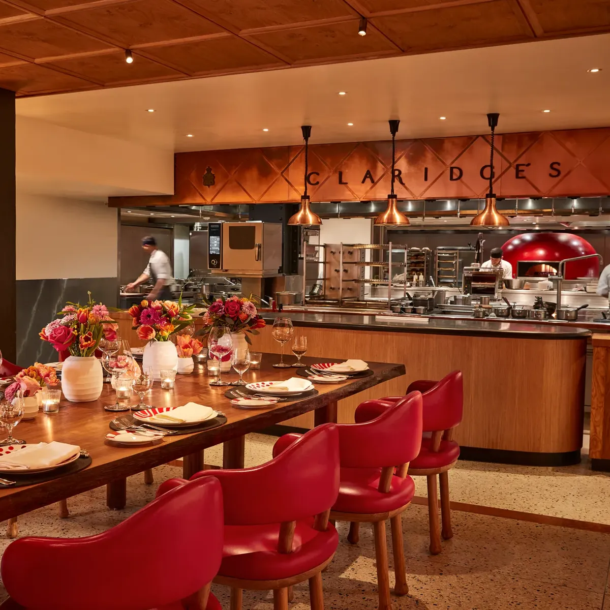 Warm, contemporary dining space with a long wooden table set with flowers, candles, and red chairs. The open kitchen in the background features copper accents, chefs at work, and “Claridge’s” displayed above the cooking area.