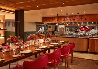 Warm, contemporary dining space with a long wooden table set with flowers, candles, and red chairs. The open kitchen in the background features copper accents, chefs at work, and “Claridge’s” displayed above the cooking area.