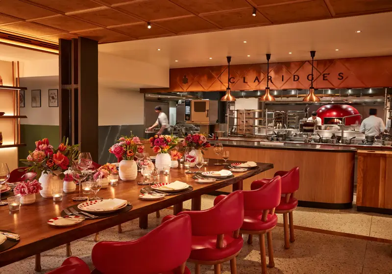 Warm, contemporary dining space with a long wooden table set with flowers, candles, and red chairs. The open kitchen in the background features copper accents, chefs at work, and “Claridge’s” displayed above the cooking area.
