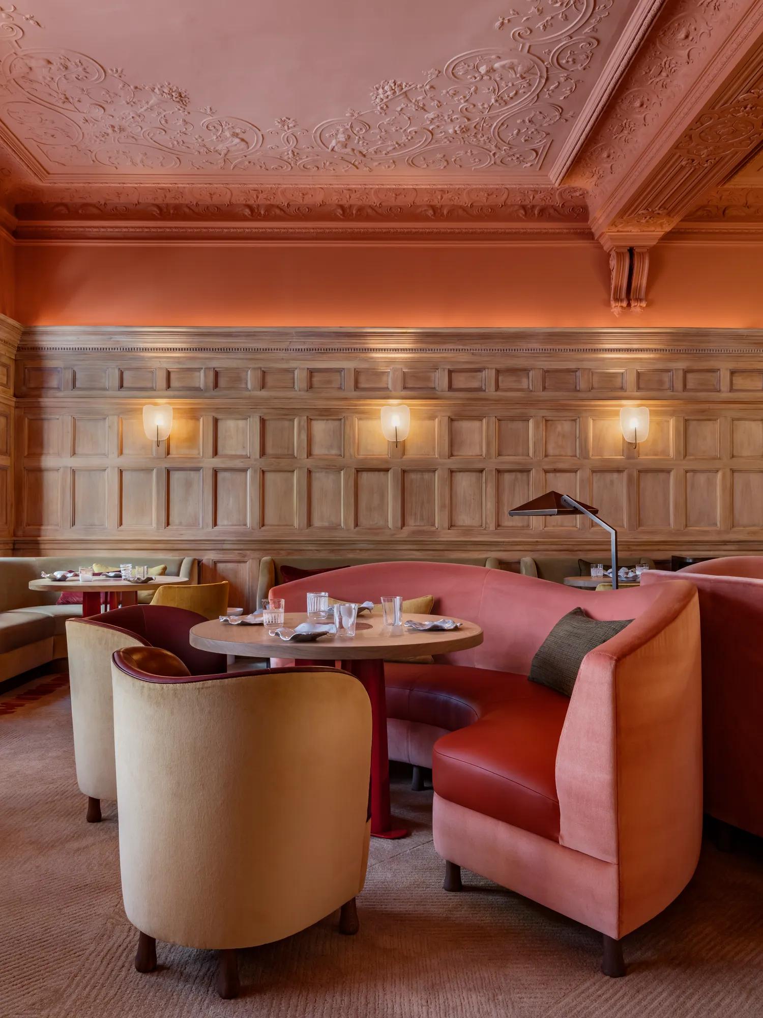 Elegant dining room with carved wood paneling, ornate ceiling, curved pink banquettes, and round tables set with glassware, under warm wall lighting.