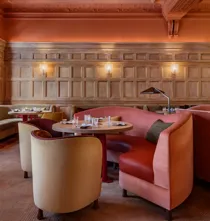 Elegant dining room with carved wood paneling, ornate ceiling, curved pink banquettes, and round tables set with glassware, under warm wall lighting.