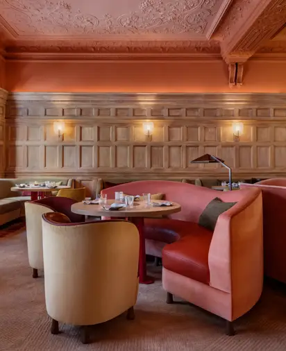 Elegant dining room with carved wood paneling, ornate ceiling, curved pink banquettes, and round tables set with glassware, under warm wall lighting.
