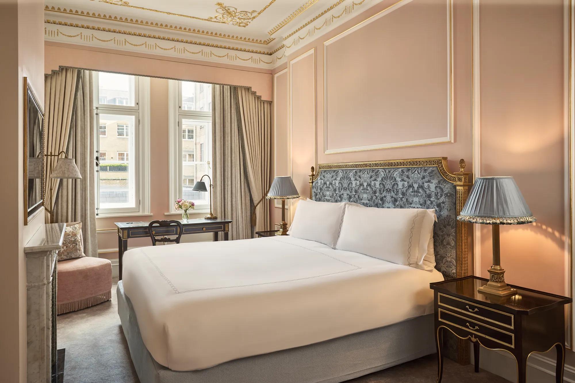 King-size bed with an upholstered headboard and bedside tables, set in a pink-toned bedroom with gold detailing, a writing desk by tall windows, and a marble fireplace.