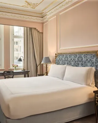 King-size bed with an upholstered headboard and bedside tables, set in a pink-toned bedroom with gold detailing, a writing desk by tall windows, and a marble fireplace.
