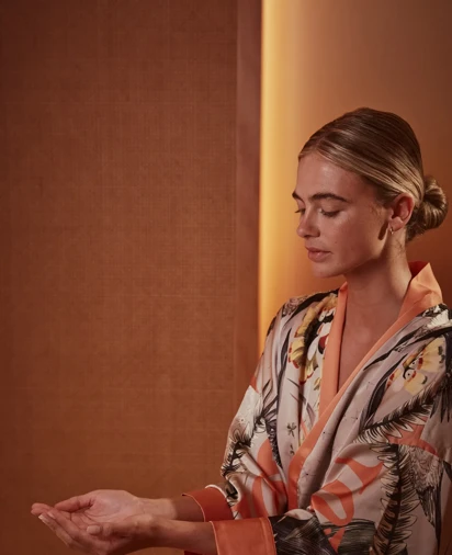 Person seated in a softly lit interior, eyes closed, holding one palm open in a calm, meditative pose, wearing a patterned robe.