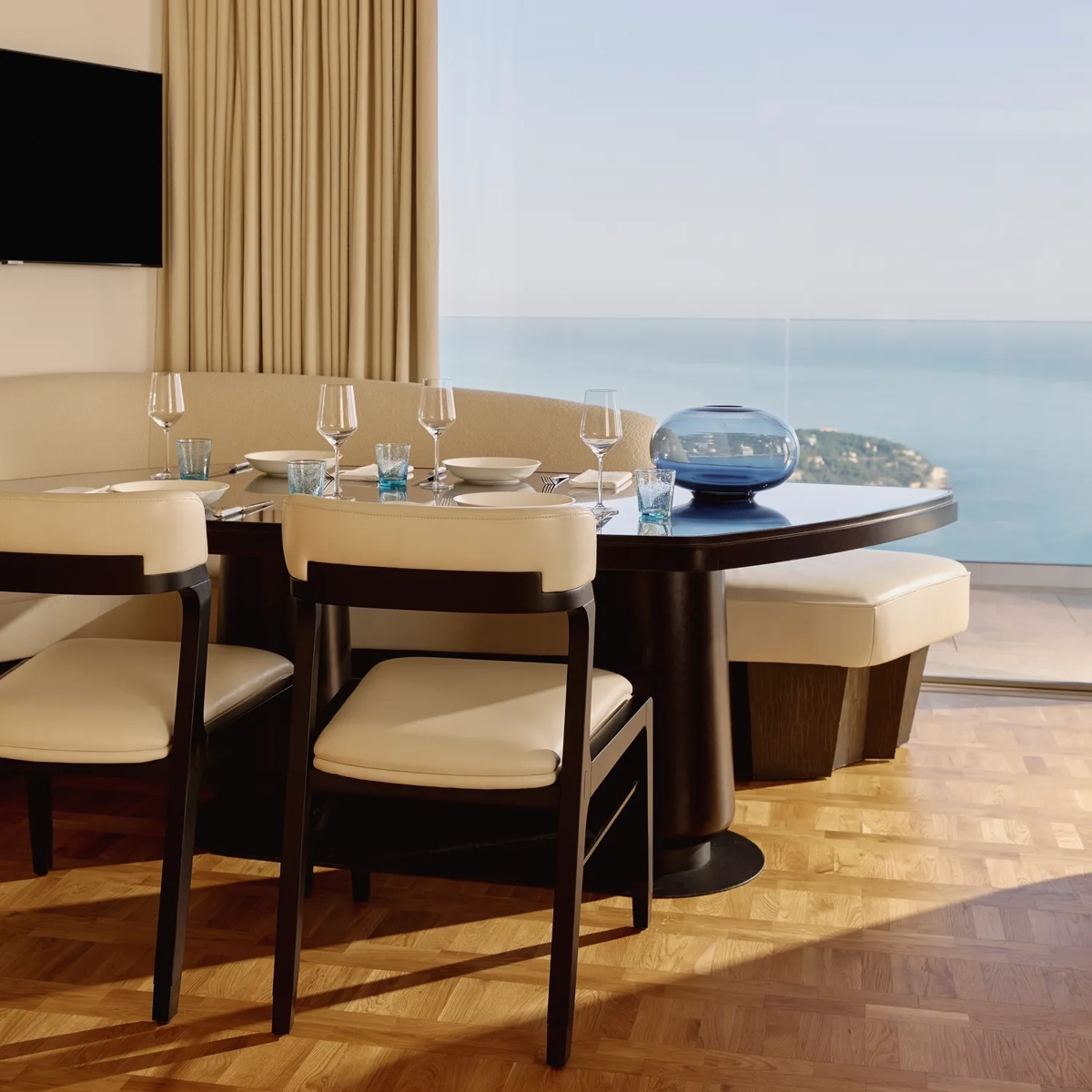 Coin repas avec table dressée, chaises rembourrées et banquette incurvée, situé près de grandes baies vitrées donnant sur la mer.