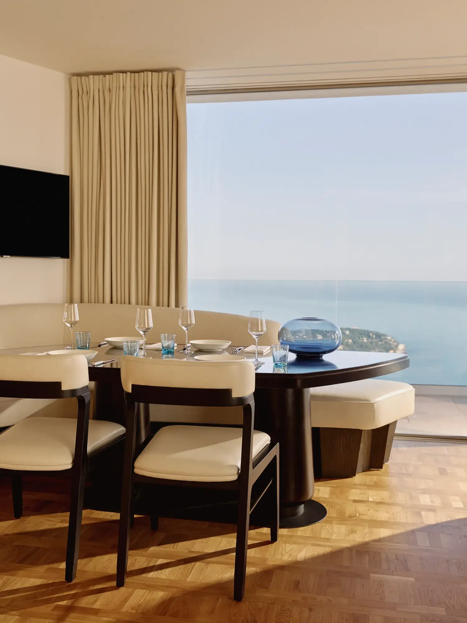 Dining area with set table, cushioned chairs, and curved banquette, positioned by large windows overlooking the sea.