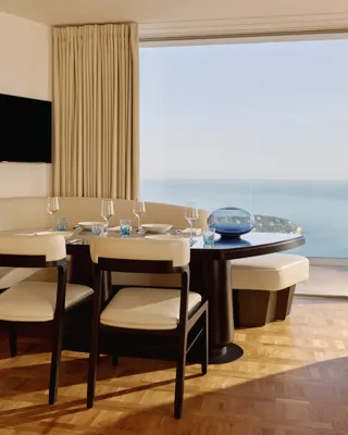 Coin repas avec table dressée, chaises rembourrées et banquette incurvée, situé près de grandes baies vitrées donnant sur la mer.