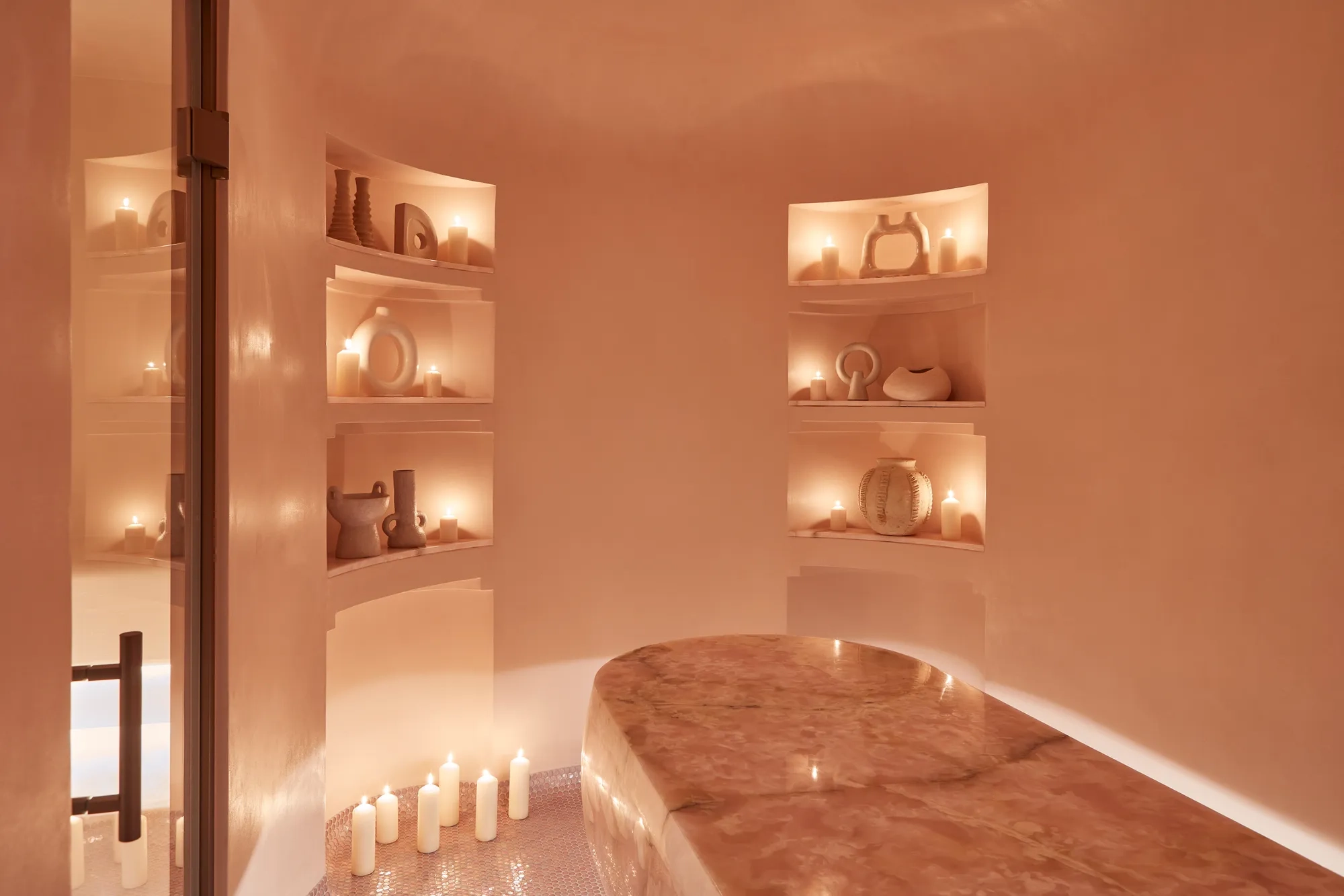 Serene spa room featuring a polished stone treatment table, softly glowing candles, and sculptural decor arranged in recessed shelves.
