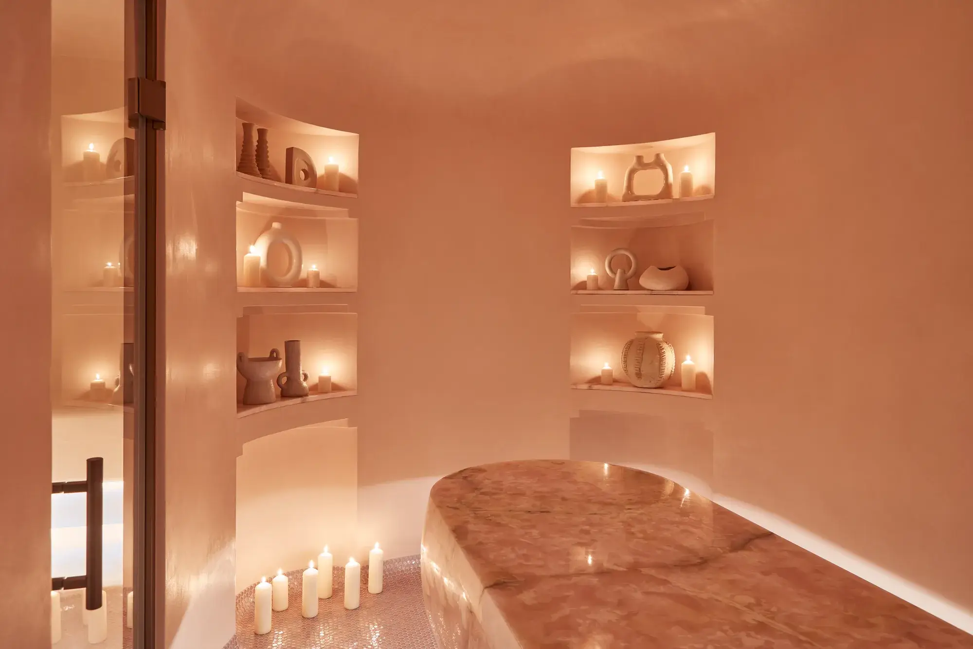 Serene spa room featuring a polished stone treatment table, softly glowing candles, and sculptural decor arranged in recessed shelves.