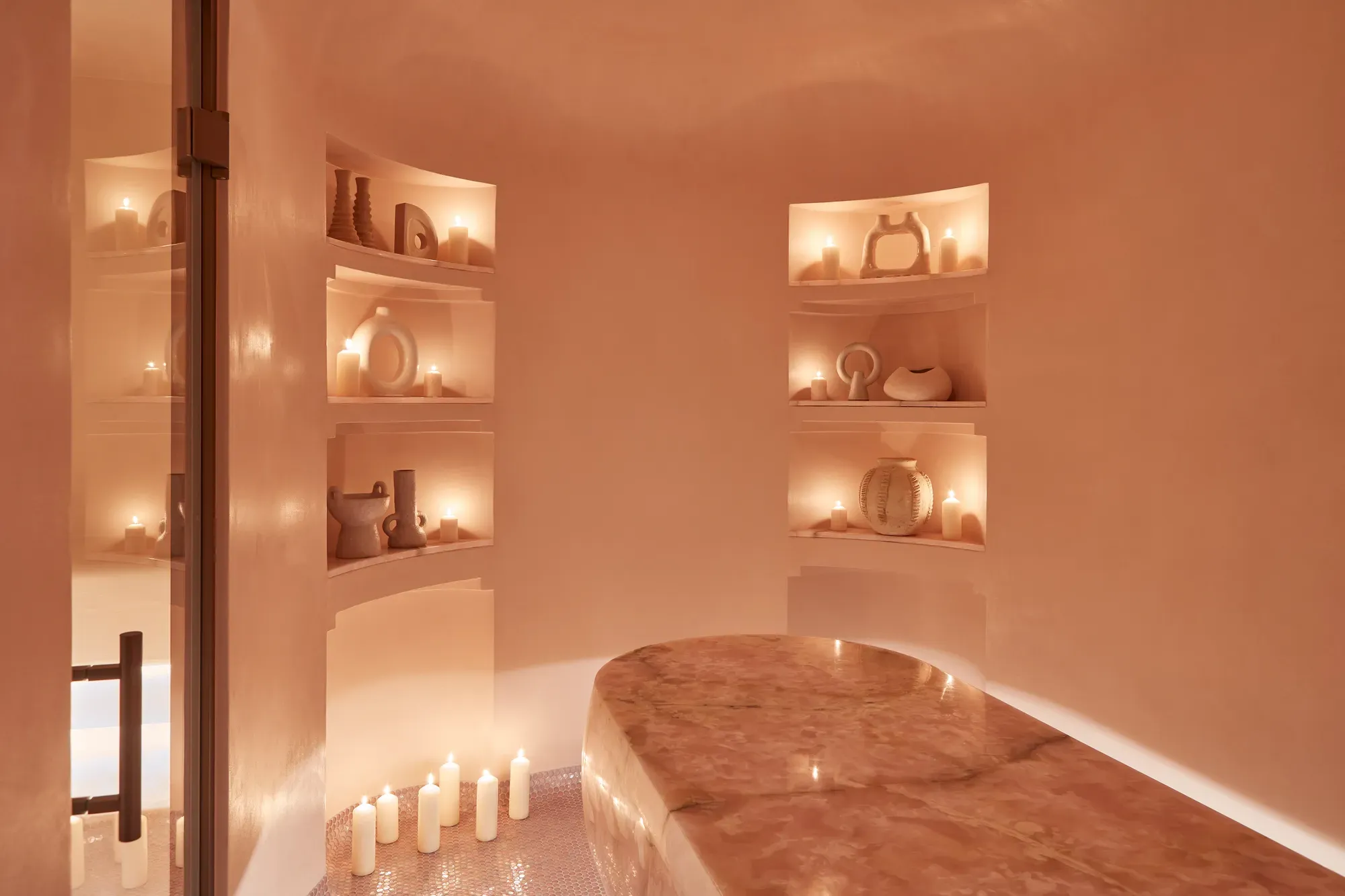 Serene spa room featuring a polished stone treatment table, softly glowing candles, and sculptural decor arranged in recessed shelves.