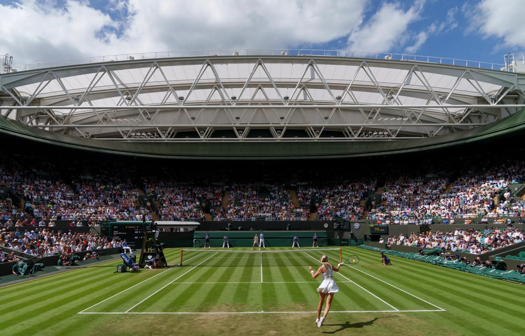 Tennis match at packed stadium, player serving on grass court under open roof and blue sky.