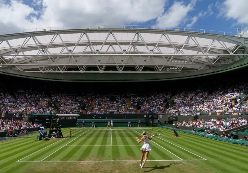 Tennis match at packed stadium, player serving on grass court under open roof and blue sky.