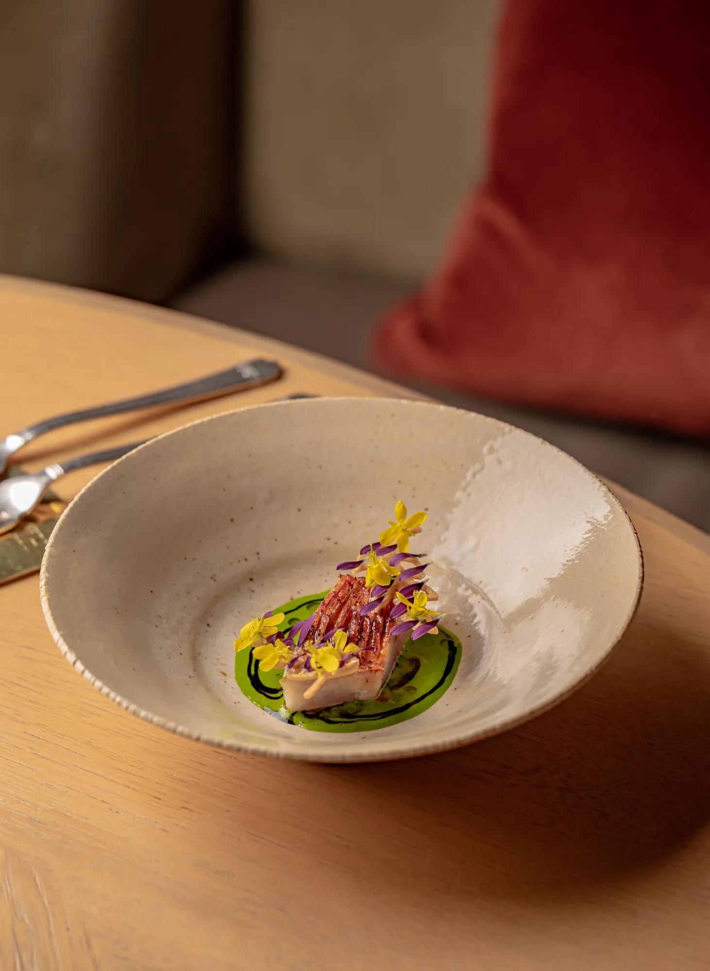 Plated gourmet dish with sliced fish, green sauce and edible flowers in a ceramic bowl on a wooden table