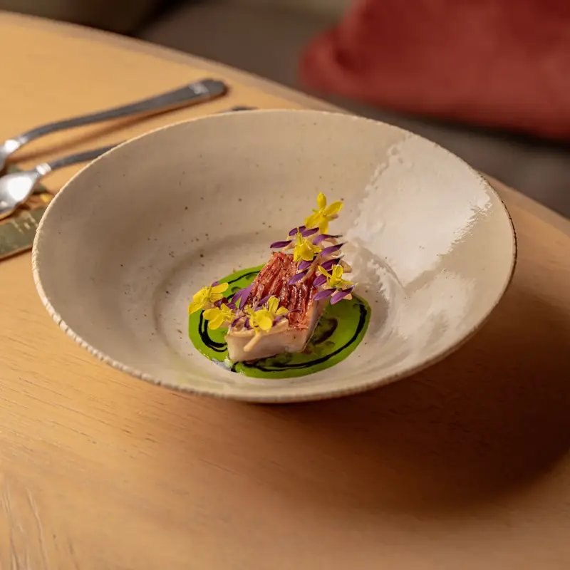Plated gourmet dish with sliced fish, green sauce and edible flowers in a ceramic bowl on a wooden table