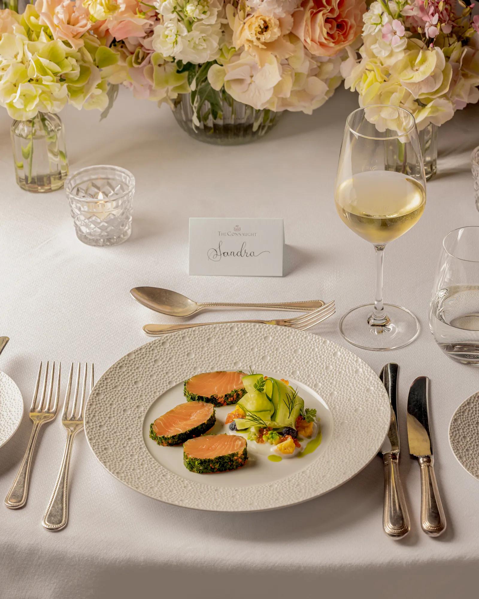Plated salmon starter with wine glass and floral centerpiece on elegant white table setting.