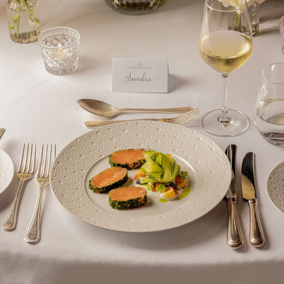 Plated salmon starter with wine glass and floral centerpiece on elegant white table setting.