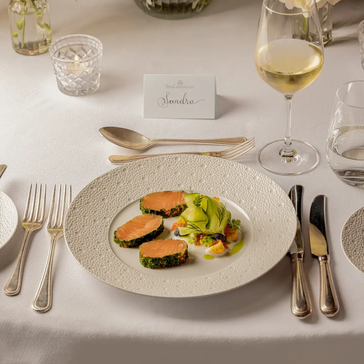 Plated salmon starter with wine glass and floral centerpiece on elegant white table setting.