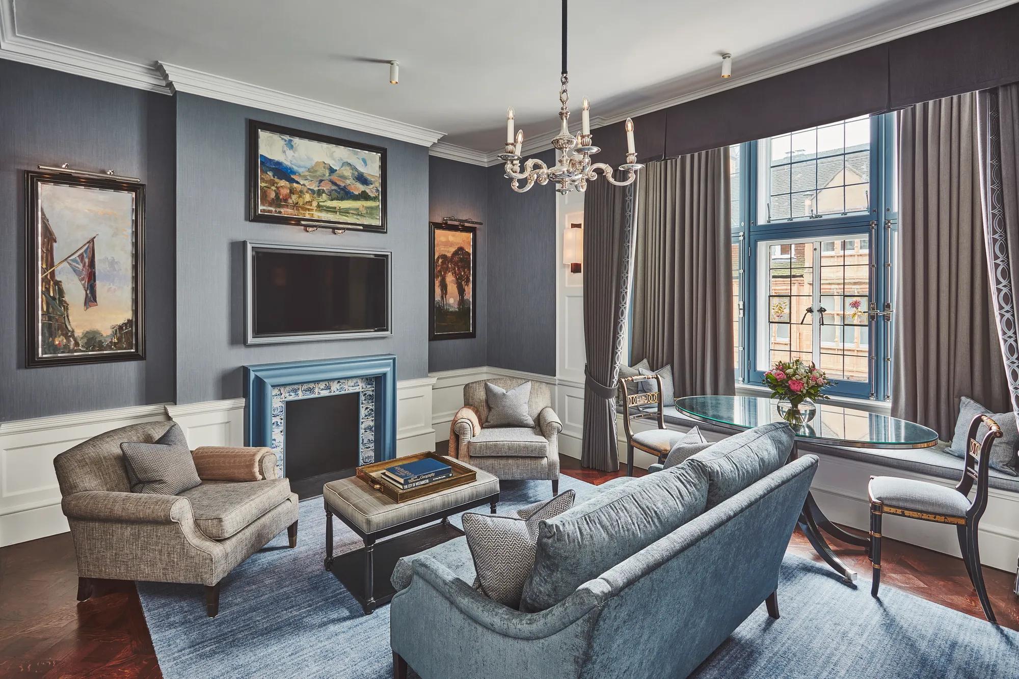 Elegant lounge with blue sofa and armchairs, fireplace, chandelier, and round dining table by large curtained window.