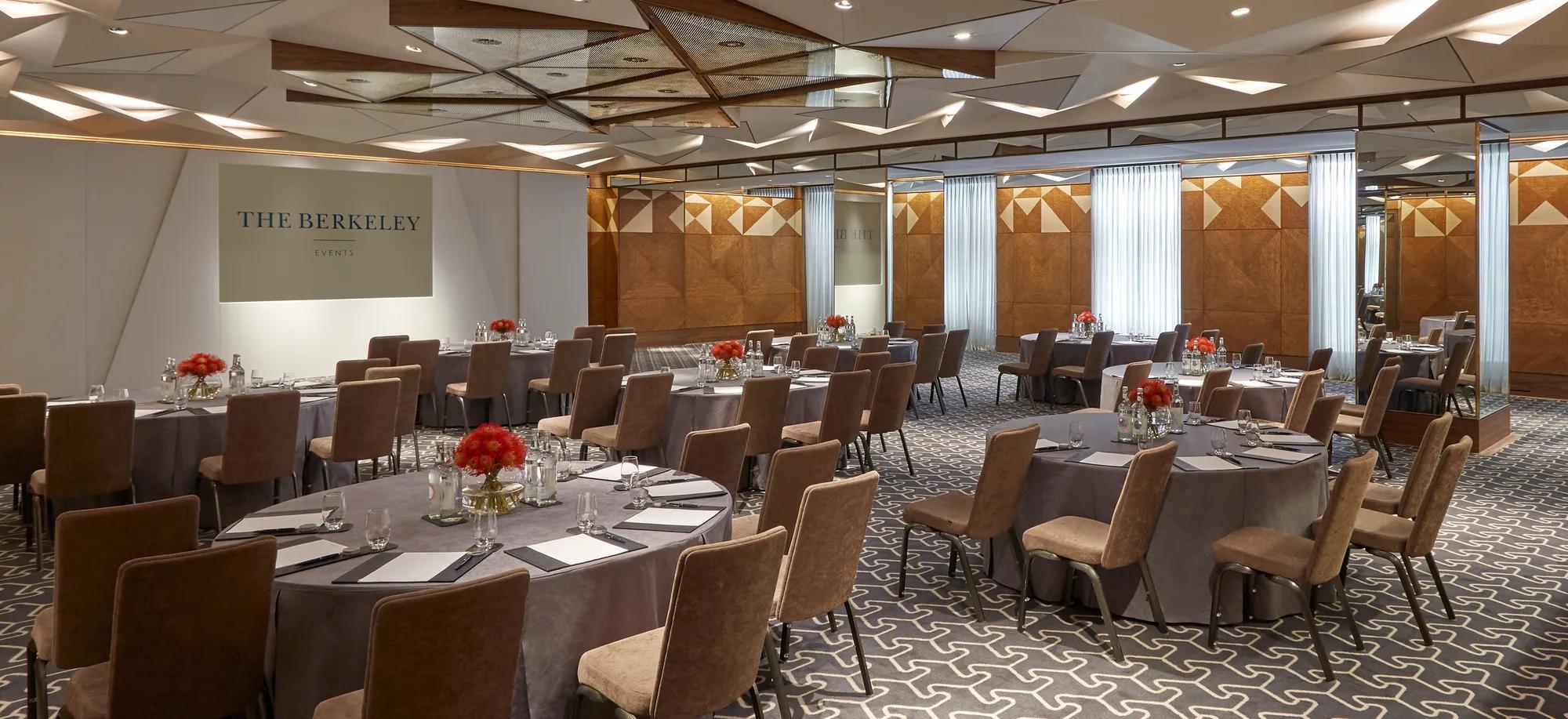 Conference room set with round tables, upholstered chairs, floral centrepieces and a presentation screen branded The Berkeley Events.