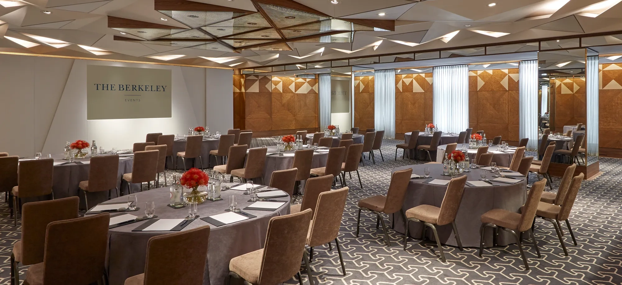 Conference room set with round tables, upholstered chairs, floral centrepieces and a presentation screen branded The Berkeley Events.